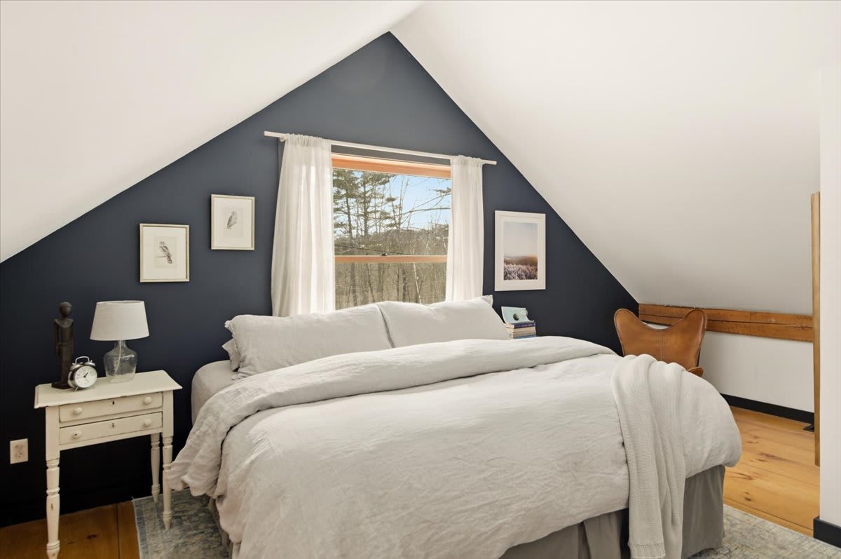 A cozy attic bedroom features a large bed with light bedding, a dark accent wall, and a window overlooking a snowy landscape. The sloped white ceiling adds architectural interest to the rustic space.