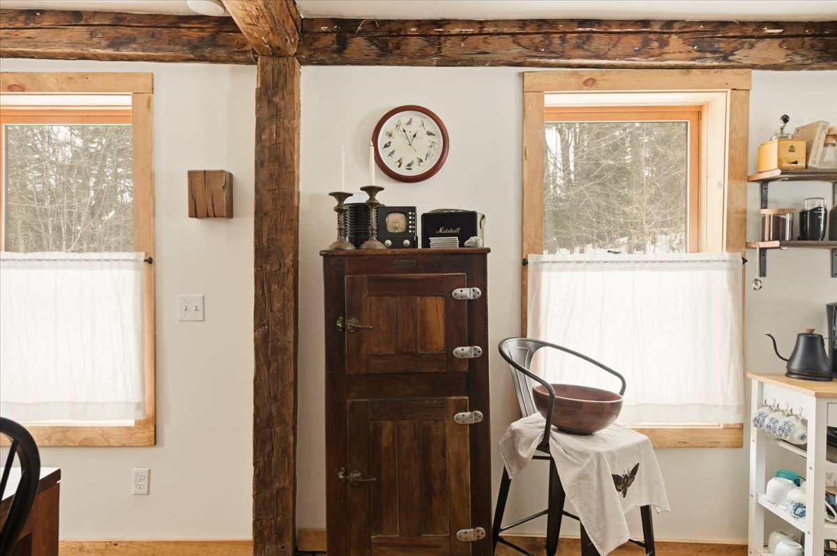 A cozy corner in the dining area, featuring a tall, dark wood vintage-style cabinet positioned between two windows with white cafe curtains. Exposed wooden beams and hardwood floors enhance the rustic charm.