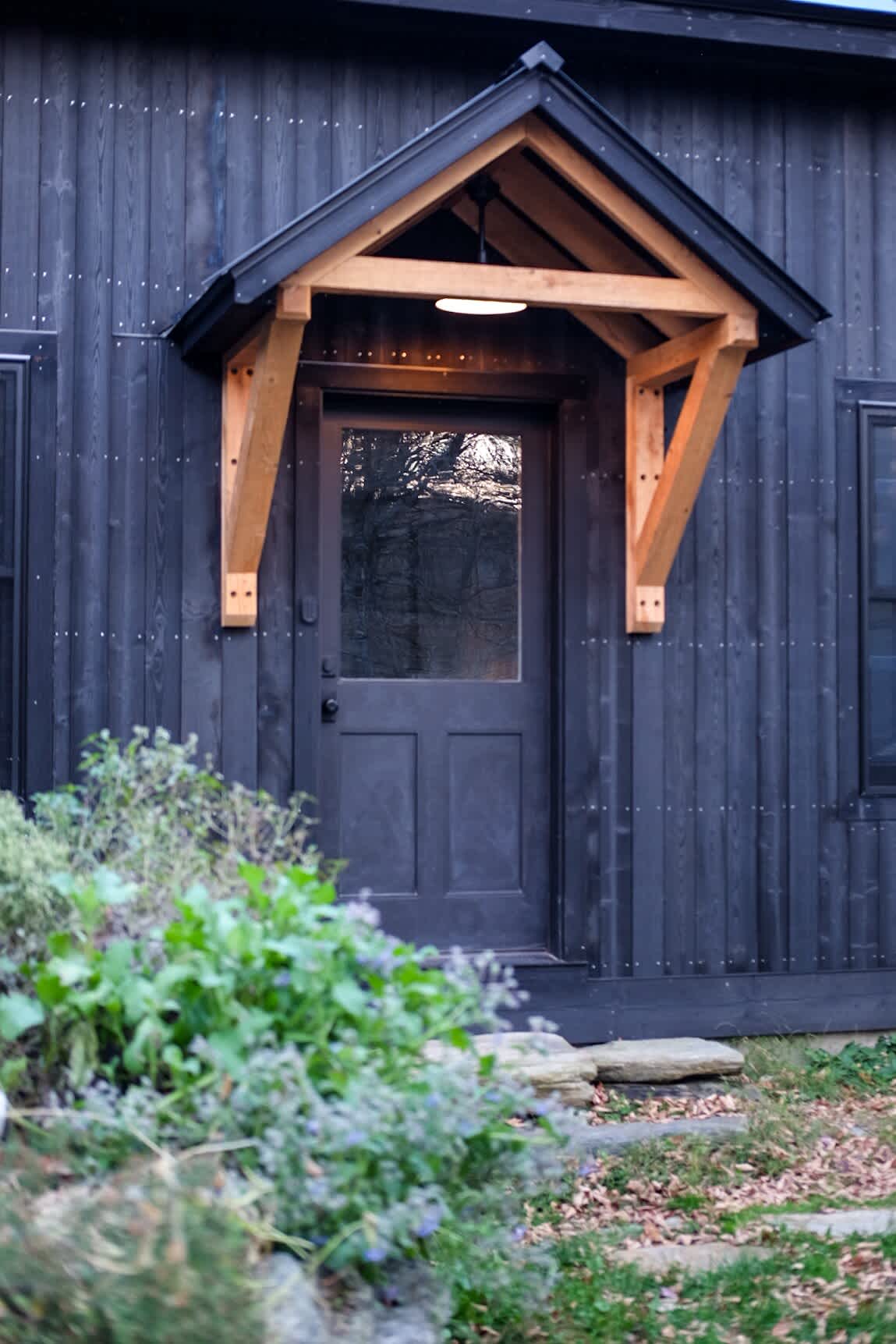 Close-up of the dark front door with a rustic wooden canopy and an overhead light, framed by lush green foliage and stone steps, suggesting a welcoming entrance.