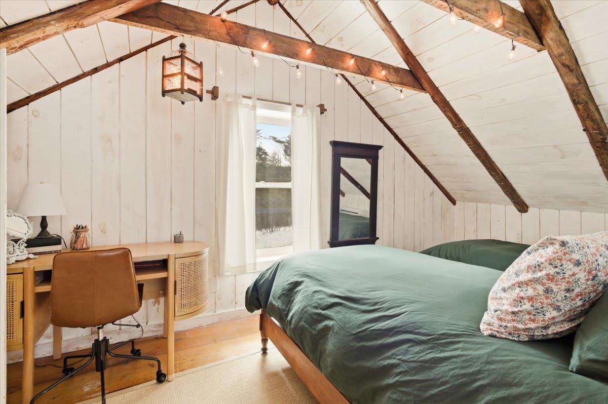 This cozy bedroom features a bed with dark green bedding, a wooden desk with a brown leather chair, and white shiplap walls, all beneath a vaulted ceiling with exposed wooden beams and string lights. A window offers natural light.
