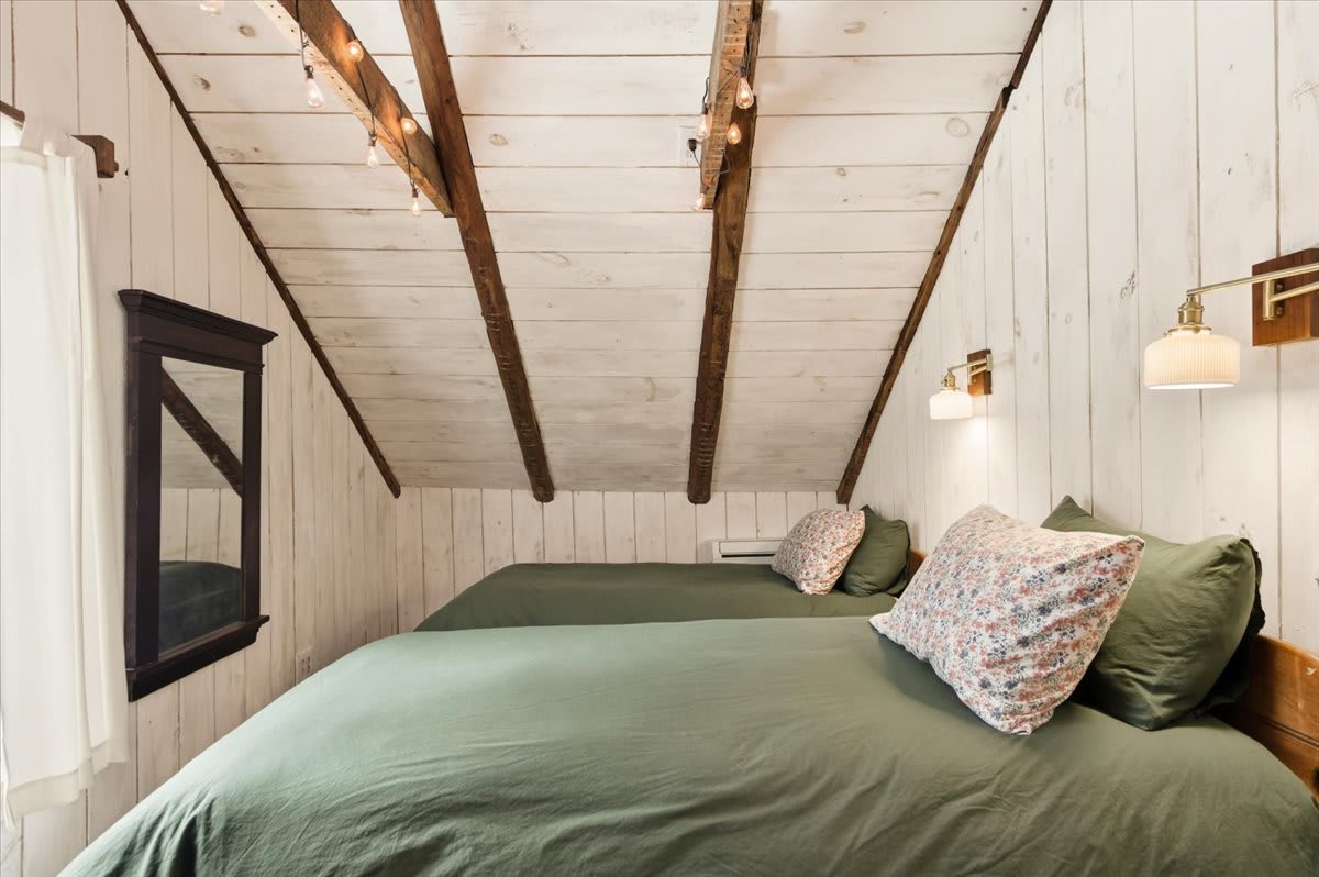 A rustic bedroom features two beds with dark green bedding and floral pillows, set against white shiplap walls, with exposed wooden beams and string lights adorning the vaulted ceiling.