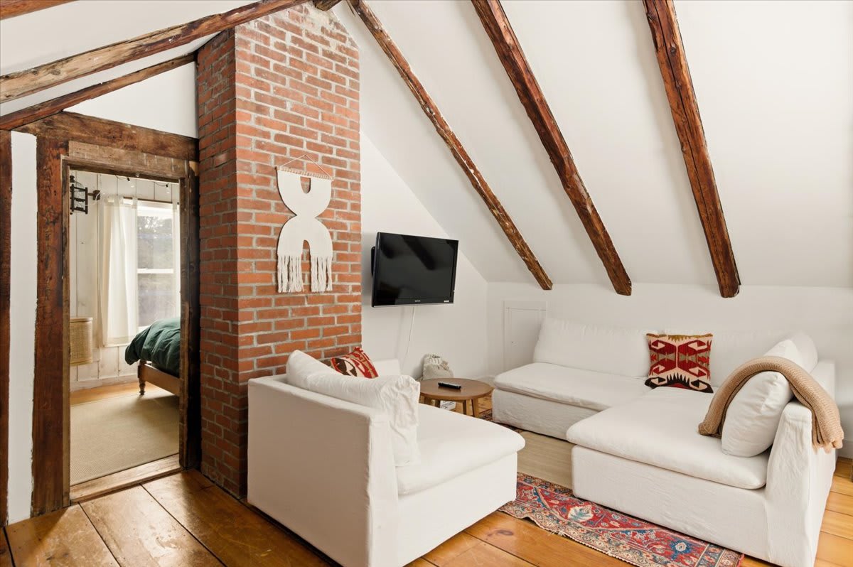 A cozy living area features white slipcovered sofas, a wall-mounted TV, and a prominent brick chimney, all beneath a vaulted ceiling with exposed wooden beams. A doorway leads to an adjacent bedroom.