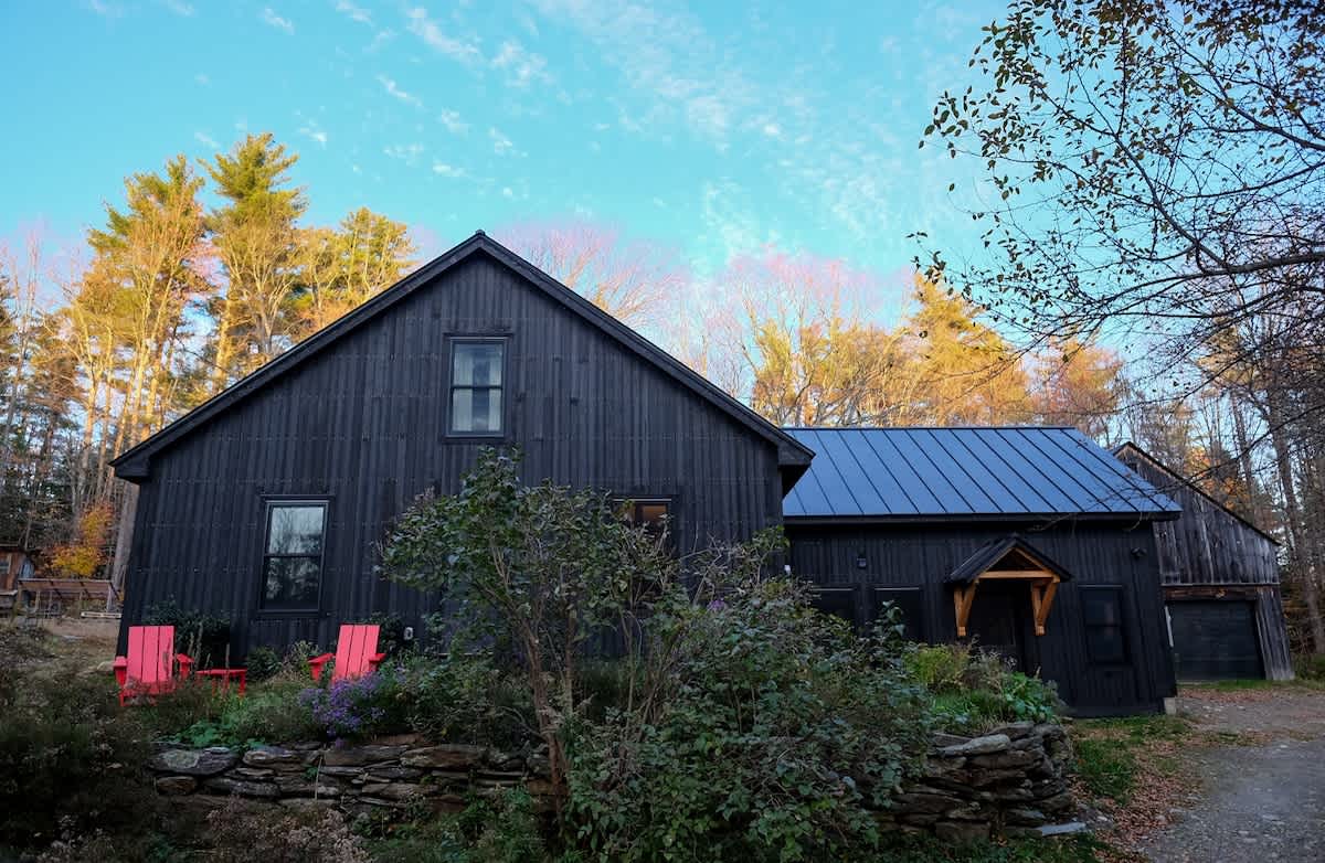 Side view of the dark-sided farmhouse with a blue metal roof, featuring a landscaped garden with red Adirondack chairs, vibrant autumn trees, and a clear blue sky.