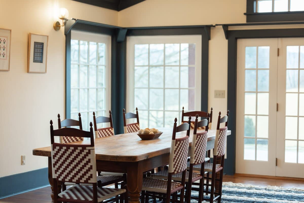A close-up of the rustic wooden dining table highlights the unique woven patterns on the backs of the eight chairs, set against a backdrop of large windows and French doors. A bowl of decorative eggs rests on the table.