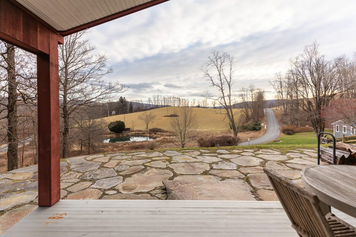 A rustic outdoor porch offers expansive views of a rolling landscape with a small pond, distant hills, and a winding road under a cloudy sky. The foreground features a stone patio and wooden deck with outdoor furniture.