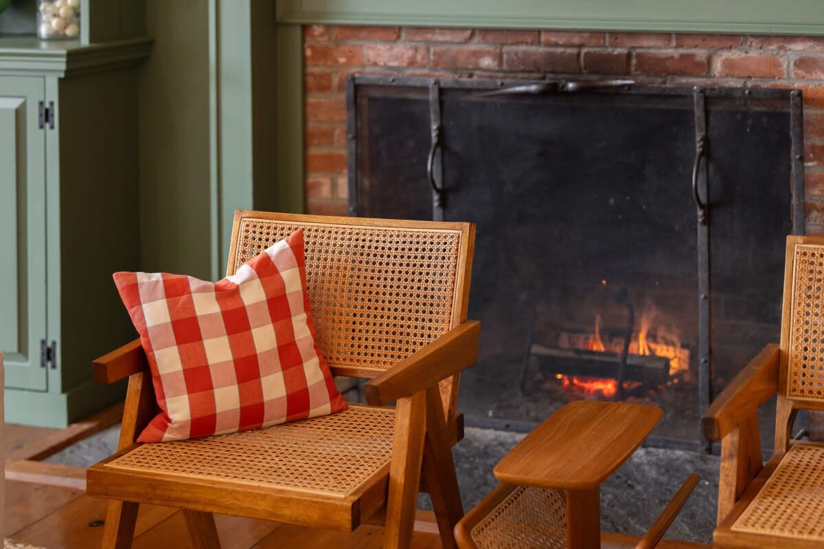 A cozy cane armchair with a red and white gingham pillow sits before a rustic brick fireplace with a black metal screen, where a warm fire glows. The scene suggests a comfortable and inviting living area.