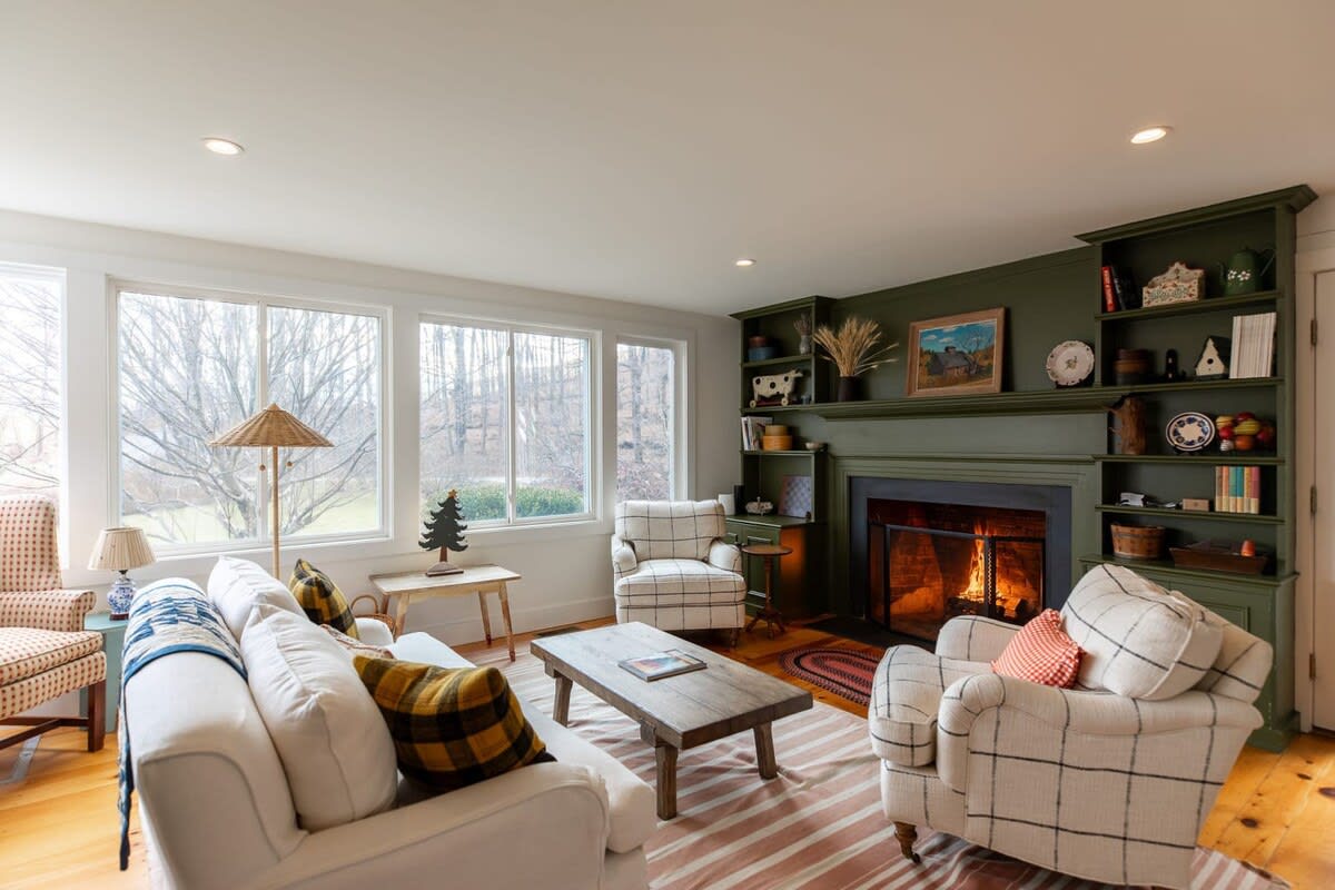 A bright living room with large windows overlooking a wooded landscape features a white sofa and two plaid armchairs arranged around a rustic wooden coffee table. A green fireplace mantel with built-in shelves adds a cozy focal point.