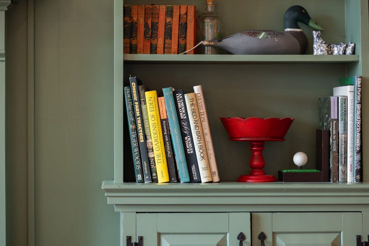 A close-up of a sage green built-in bookshelf filled with books and decorative items, including a wooden duck decoy and a red pedestal bowl. The shelves are painted a muted green, complementing the rustic decor.