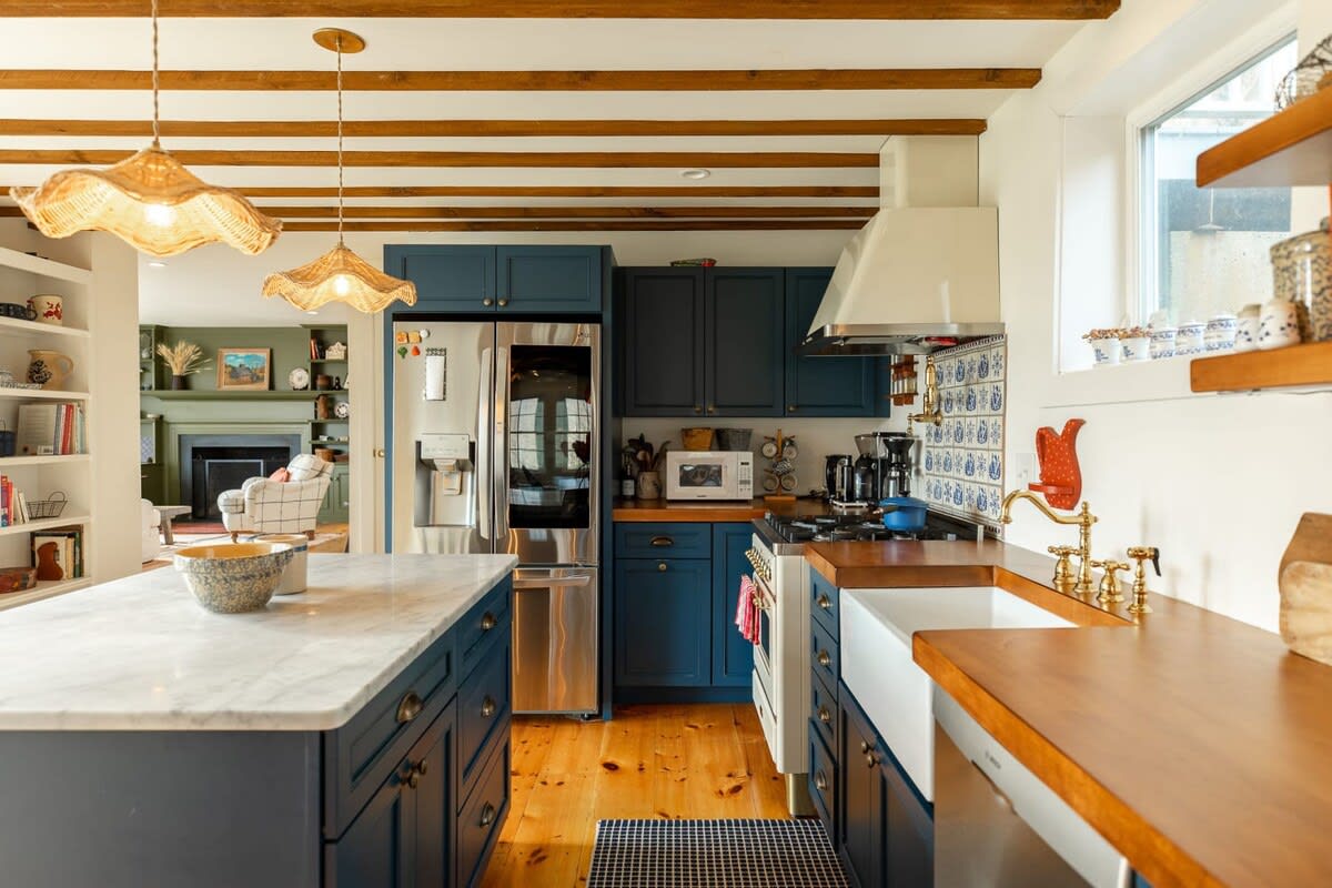 A rustic kitchen features navy blue shaker cabinets, wooden beamed ceilings, and light wood floors. It includes a white marble island, stainless steel appliances, and a white farmhouse sink with brass fixtures and a blue and white tiled backsplash.