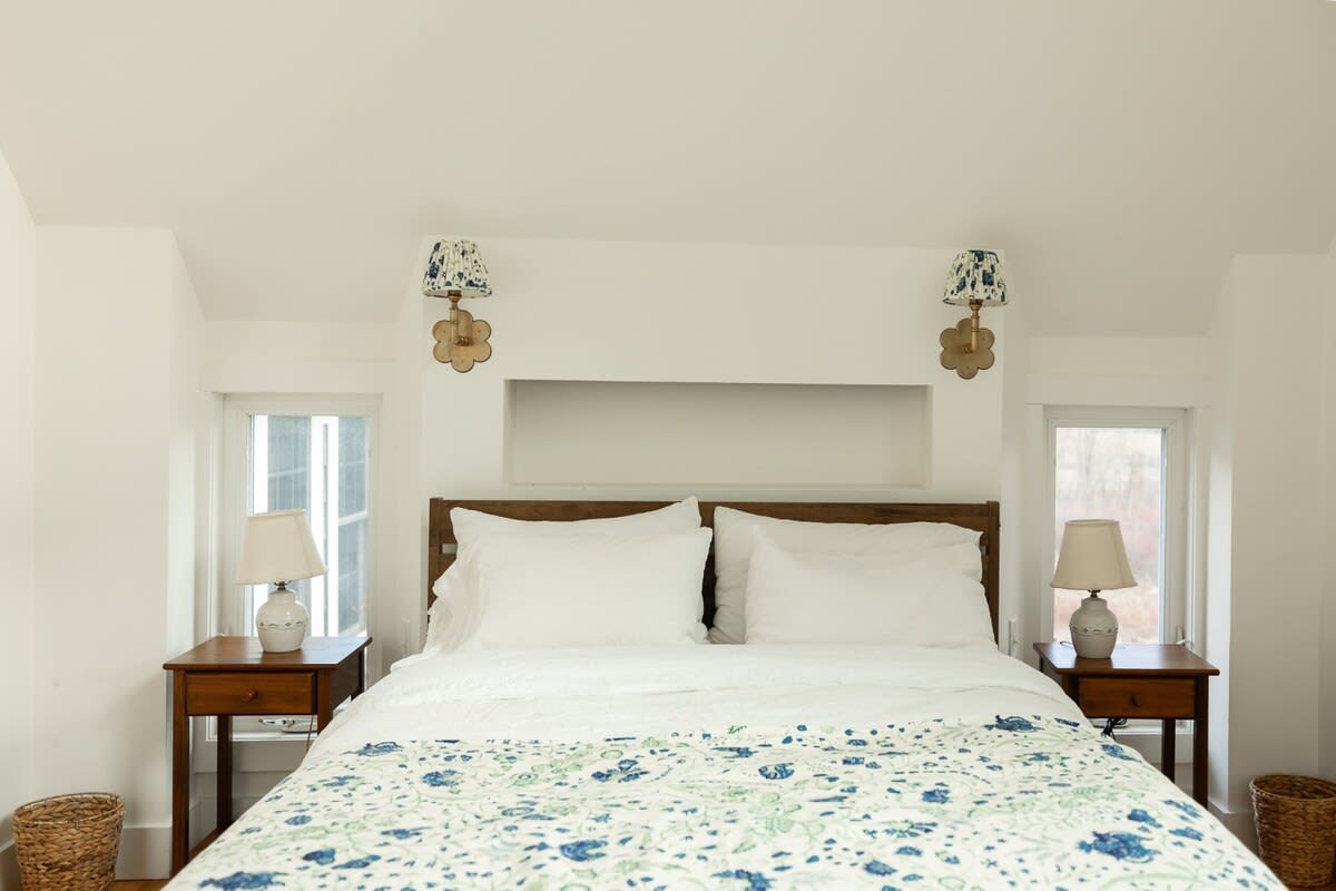 Bright bedroom featuring a wooden headboard bed with white bedding and a blue and green patterned duvet. Two wooden nightstands with ceramic lamps flank the bed, complemented by floral wall sconces above.