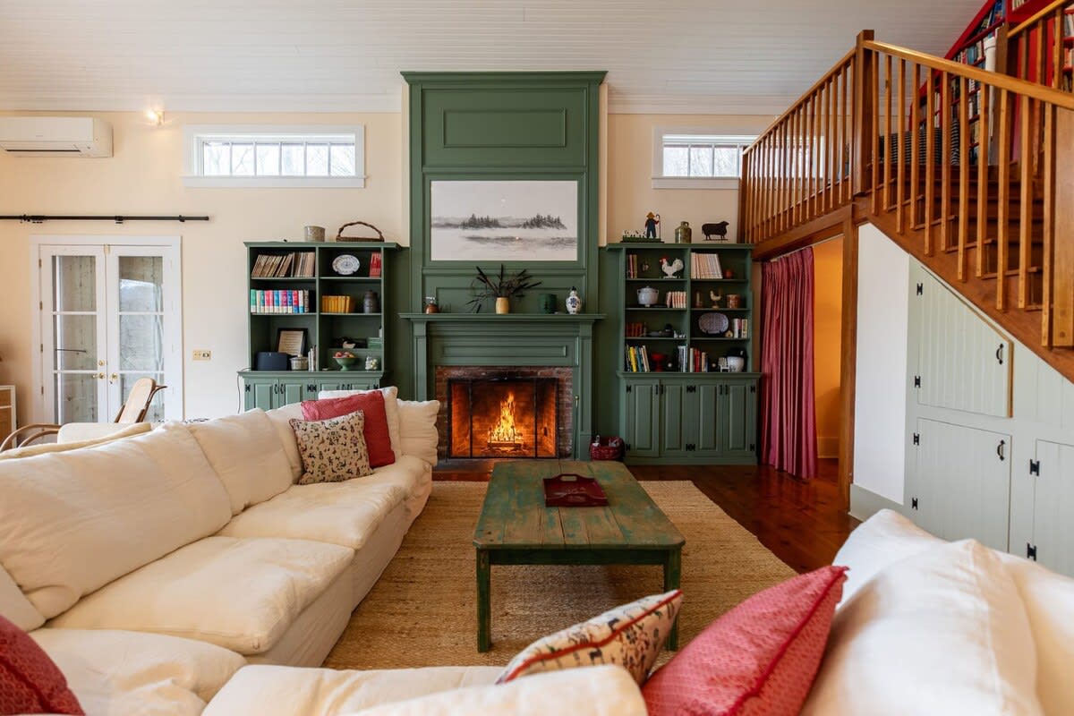 A spacious living room with a high ceiling features a large white sectional sofa facing a brick fireplace with a green mantel and built-in bookshelves. A rustic green coffee table sits on a natural fiber rug, with a wooden staircase leading to an upper level.