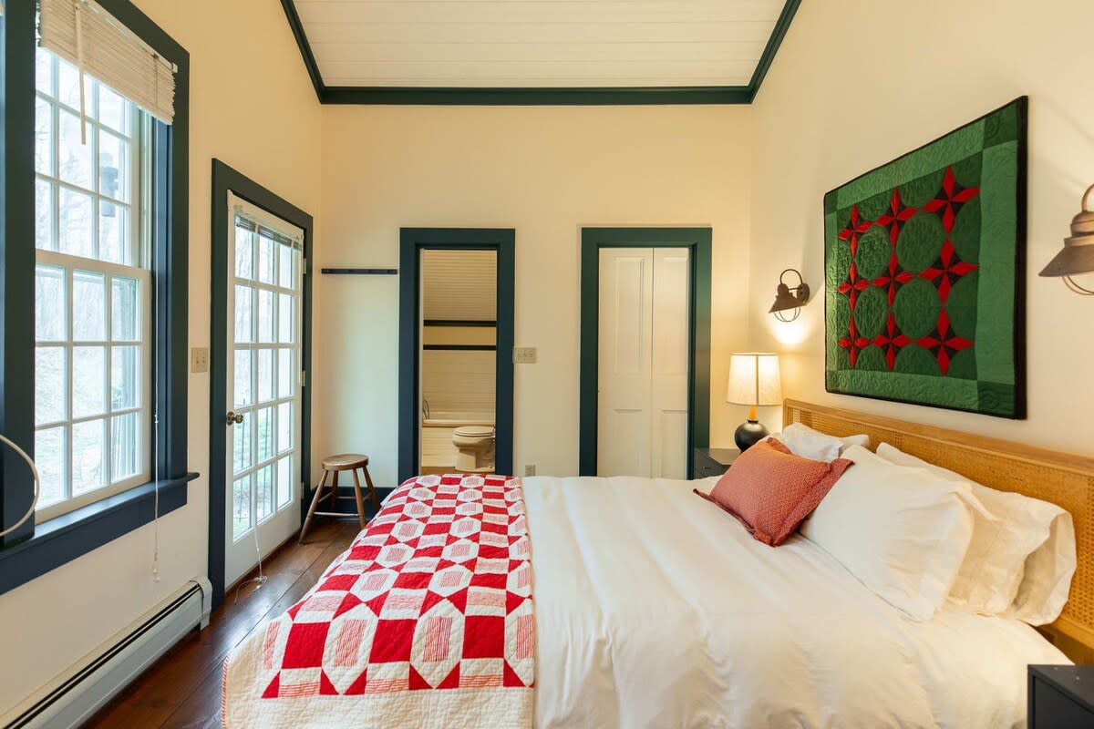 A cozy bedroom features a bed with white linens and a red and white patterned quilt, dark trim around windows and doors, and a green and red quilted wall hanging. A glimpse into a bathroom with a tub is visible through an open door.