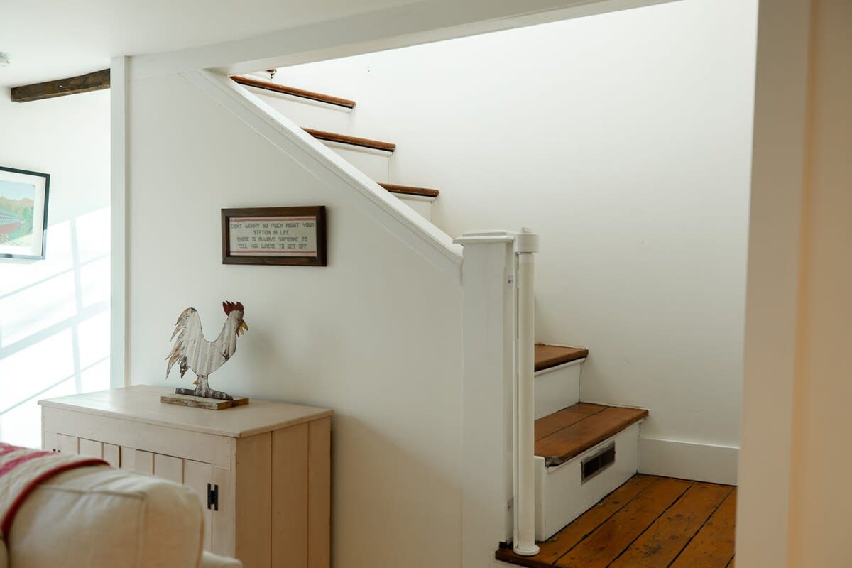 A bright hallway features a wooden staircase with white risers and banister, light wood floors, and white walls. A decorative rooster statue sits on a small white cabinet, and a framed sign hangs on the wall.