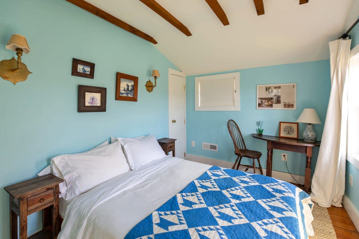 A serene bedroom features light blue walls, exposed wooden ceiling beams, and light wood floors. It includes a bed with a white comforter and a blue and white patterned quilt, flanked by rustic wooden nightstands.
