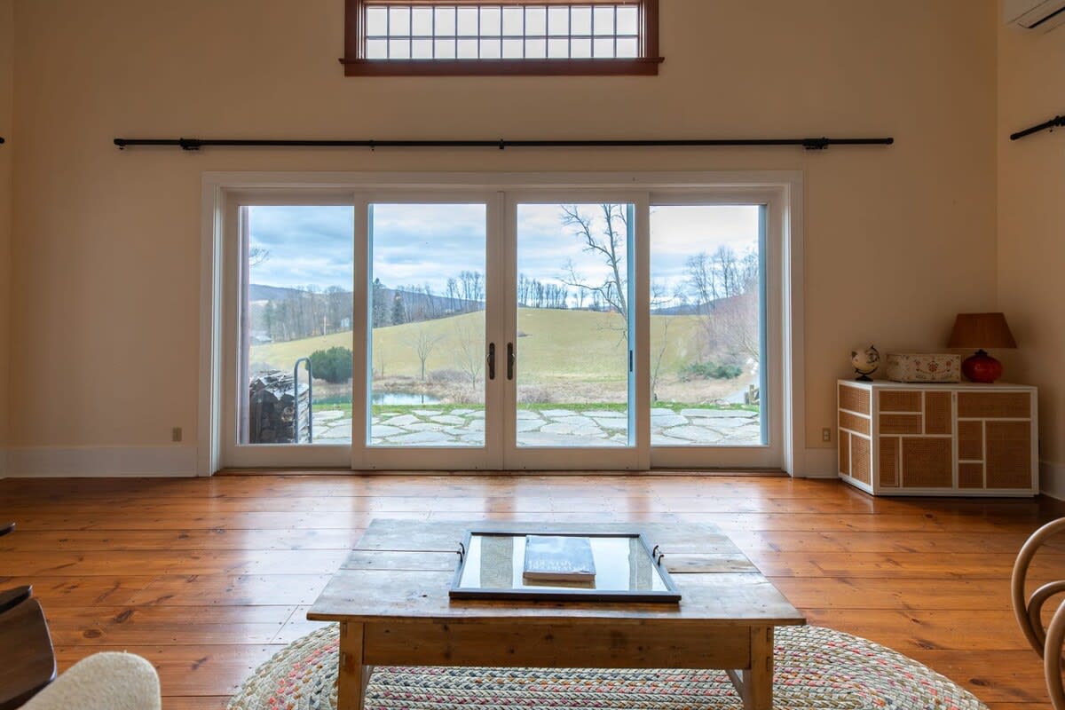 A spacious living room features wide-plank hardwood floors and large sliding glass doors offering panoramic views of rolling green hills and a tranquil pond. A rustic wooden coffee table anchors the center of the room.