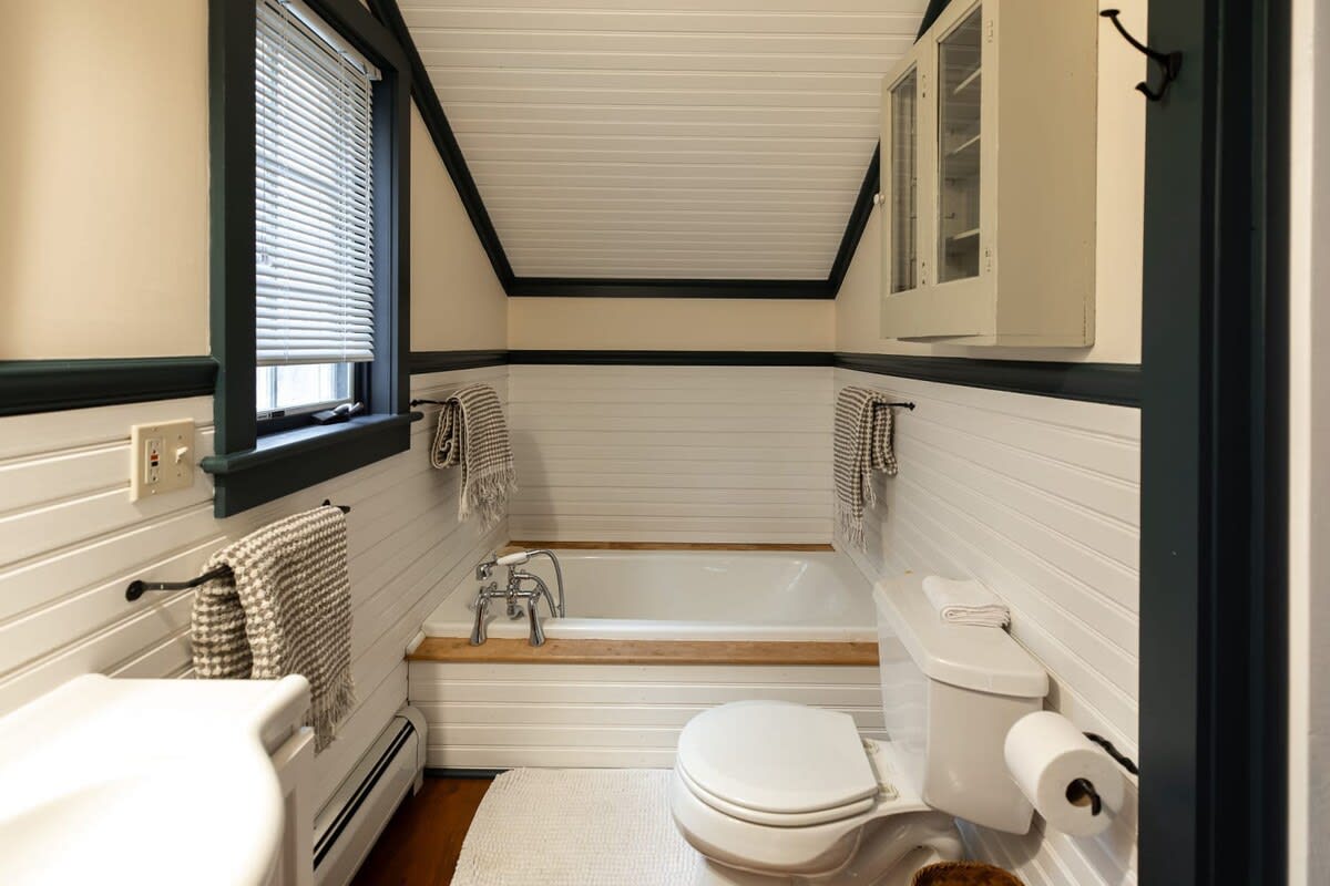 A compact bathroom features white beadboard wainscoting and a built-in bathtub with a wooden surround, alongside a toilet and pedestal sink. Dark trim frames the window and ceiling, complementing the rustic aesthetic.