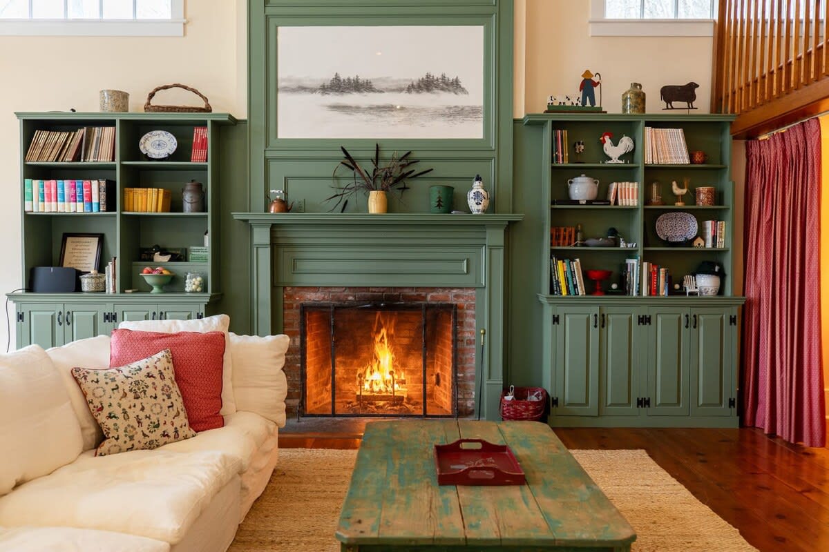 A cozy living room features a large white sectional sofa facing a brick fireplace with a roaring fire, flanked by built-in green bookshelves. A rustic green coffee table sits on a natural fiber rug, with a wooden staircase visible to the right.