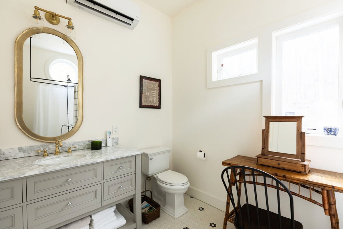 Bright bathroom featuring a gray vanity with a marble countertop and a large gold-framed oval mirror. A toilet is positioned next to the vanity, and a bamboo console table with a small wooden mirror sits under a window.