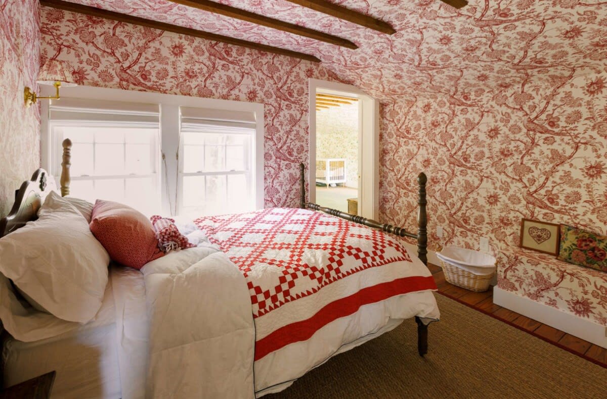 A charming bedroom features red and white floral patterned wallpaper covering the walls and ceiling, exposed wooden beams, and a dark wooden four-poster bed with red and white quilted bedding.