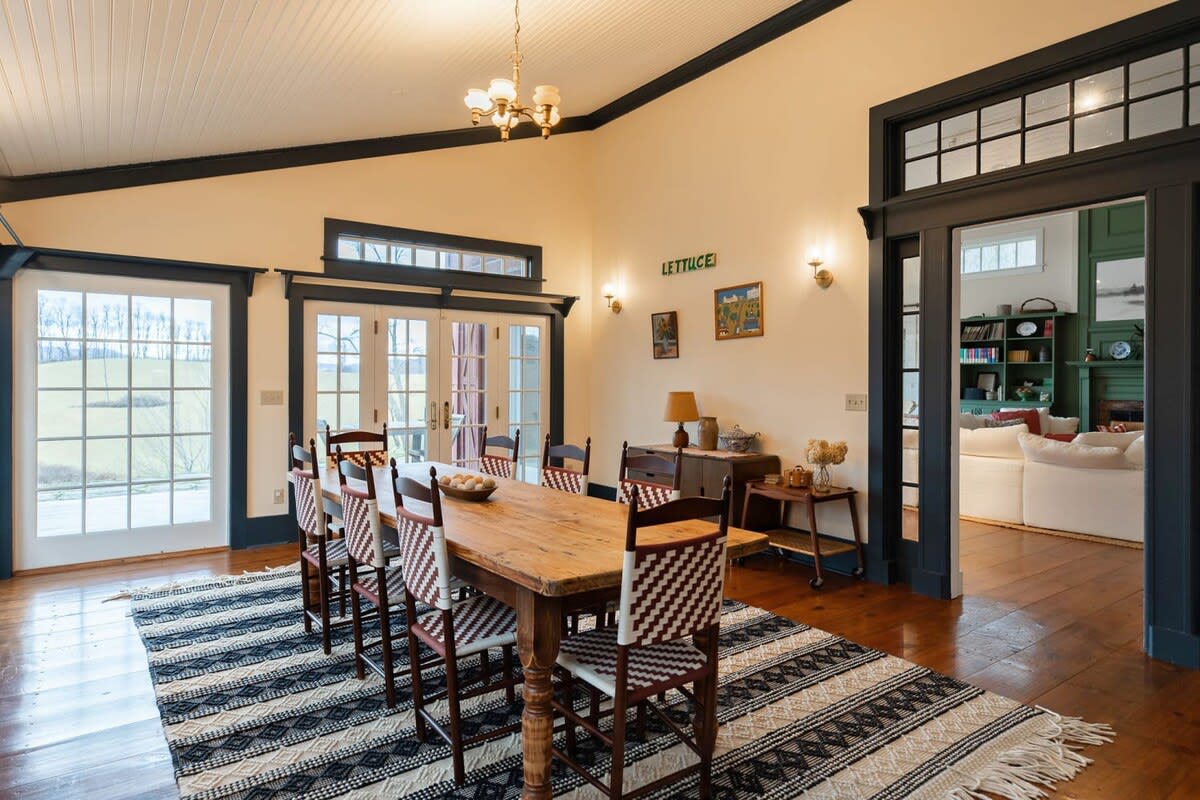 A rustic wooden dining table with eight chairs featuring woven backs is centered on a striped rug, flanked by large French doors and windows offering outdoor views. Dark trim frames the doorways, leading to other areas of the home.