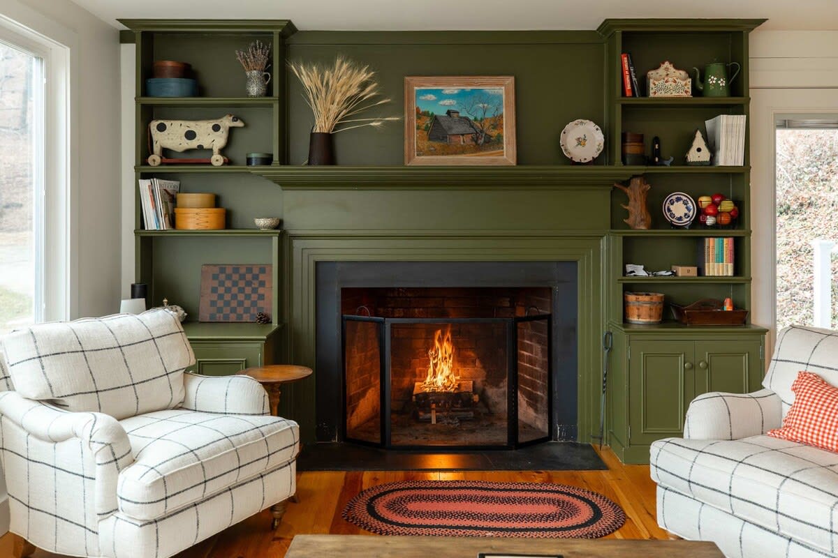 A charming living room centers around a large, olive-green fireplace with built-in shelving adorned with decorative items. Two plaid armchairs face the crackling fire, creating a warm and inviting atmosphere.