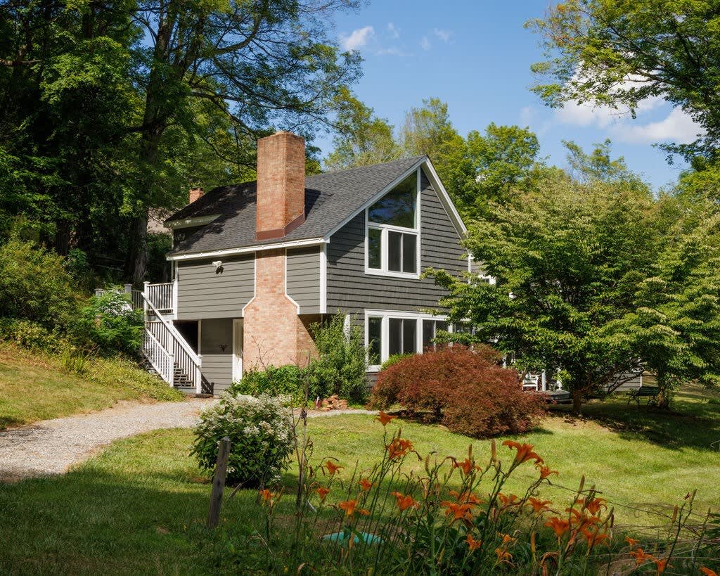 A charming gray house with a dark roof and a prominent brick chimney stands amidst lush greenery and trees. A gravel path leads up to the house, with vibrant orange daylilies blooming in the foreground.