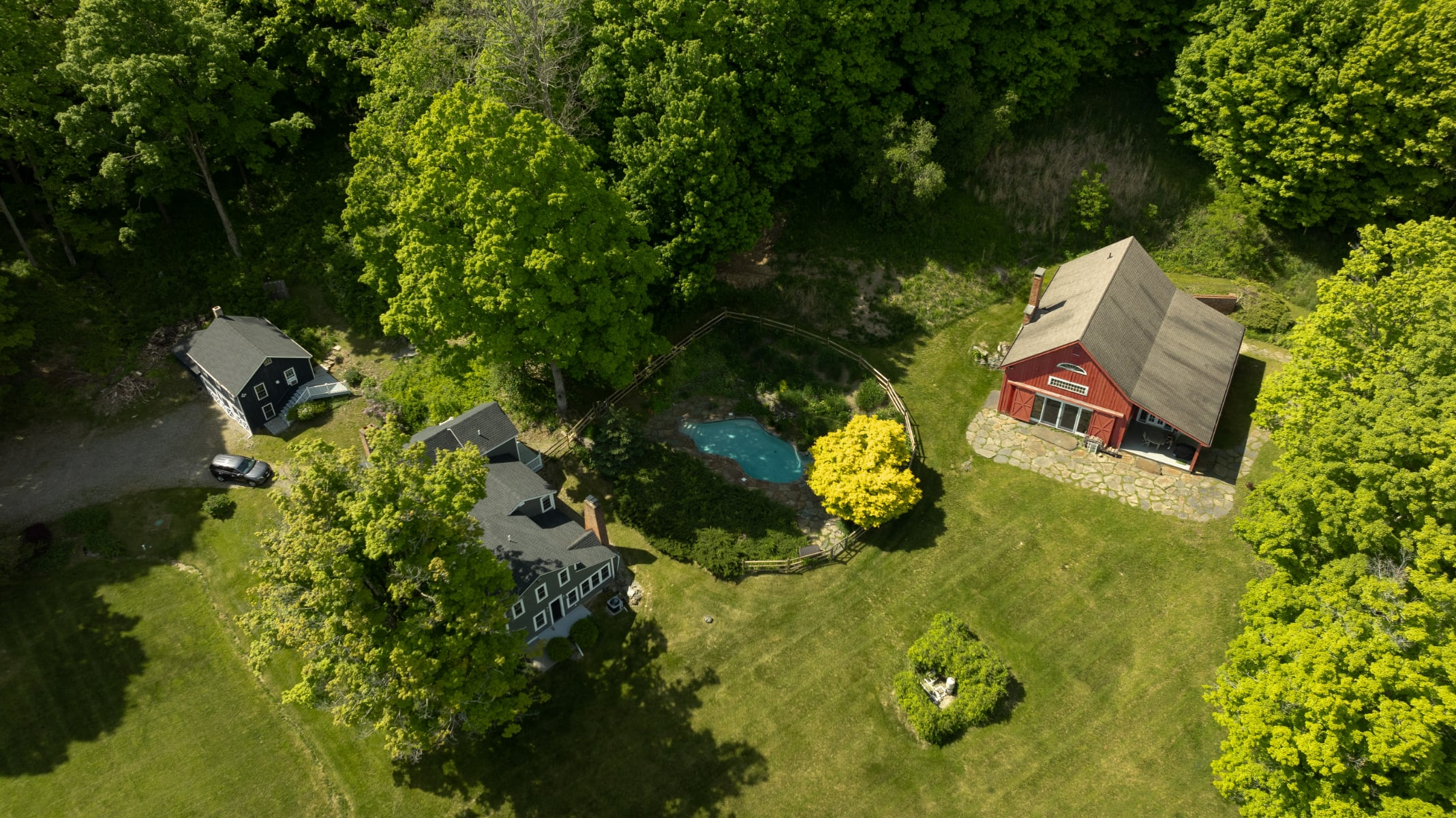 Aerial view of the estate showing a gray main house, a dark gray guesthouse, and a red barn-like structure. A fenced-in pool with a blue bottom is visible between the main house and the red barn, all set within a lush green landscape.