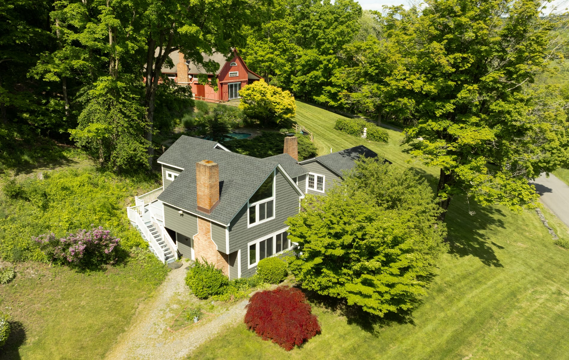 Aerial view of a gray multi-story house with a dark roof and brick chimney, nestled among lush green trees. A red barn-like building is visible further up the hill, with a glimpse of a pool.