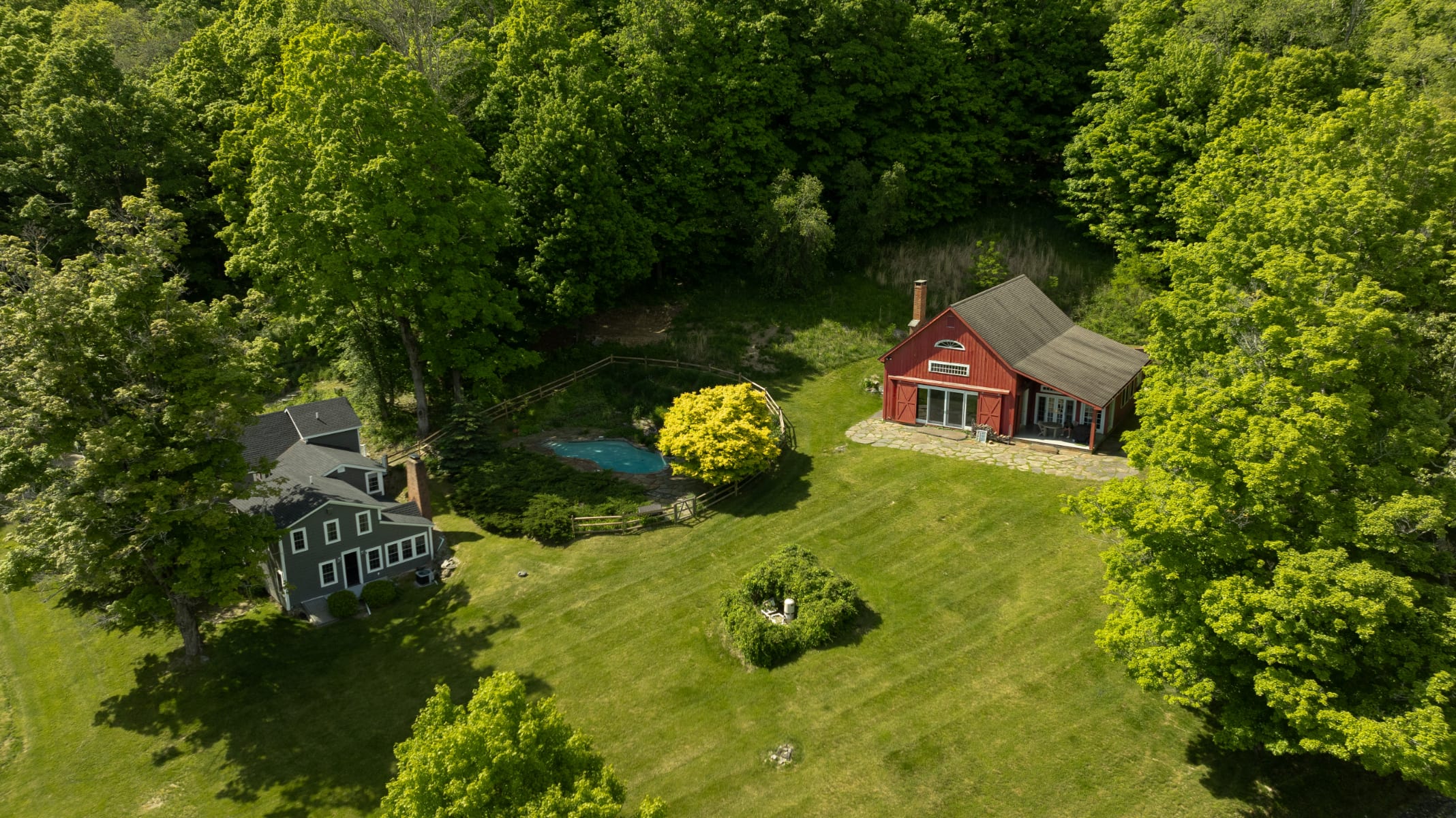 An aerial view reveals two houses on a sprawling green property surrounded by lush trees. A red cottage is on the right, and a smaller gray house is on the left, with a freeform pool and a bright yellow tree between them.