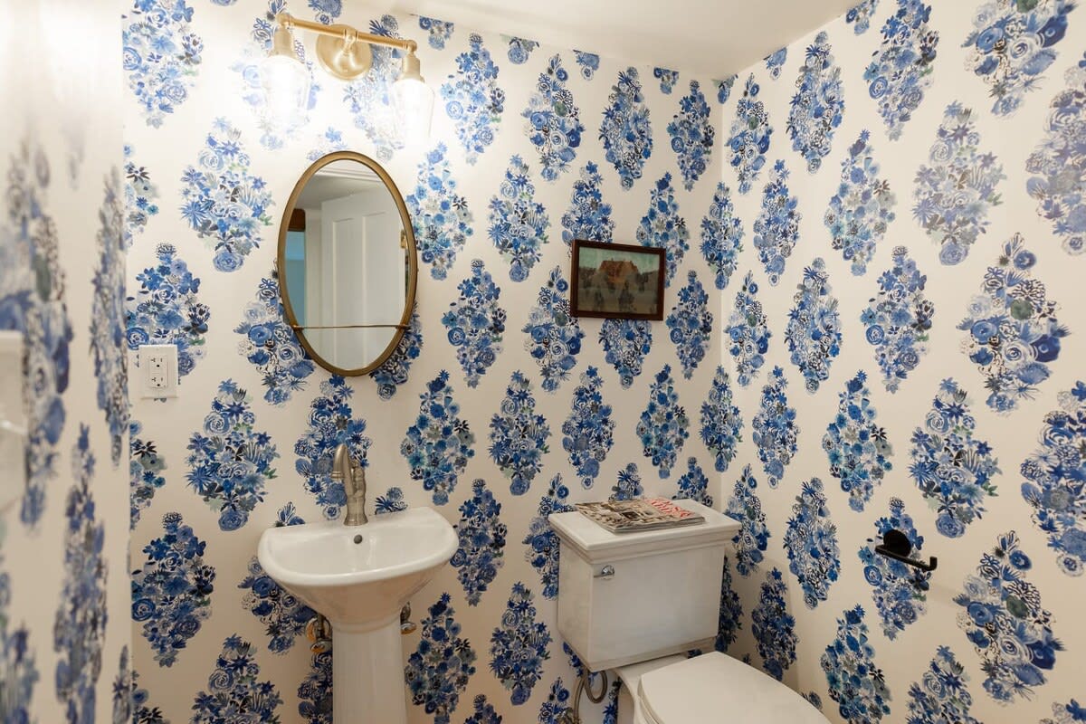 A small powder room features bold blue and white floral wallpaper, a white pedestal sink with a gold-framed oval mirror, and a white toilet. A small framed picture hangs on the wall above the toilet.