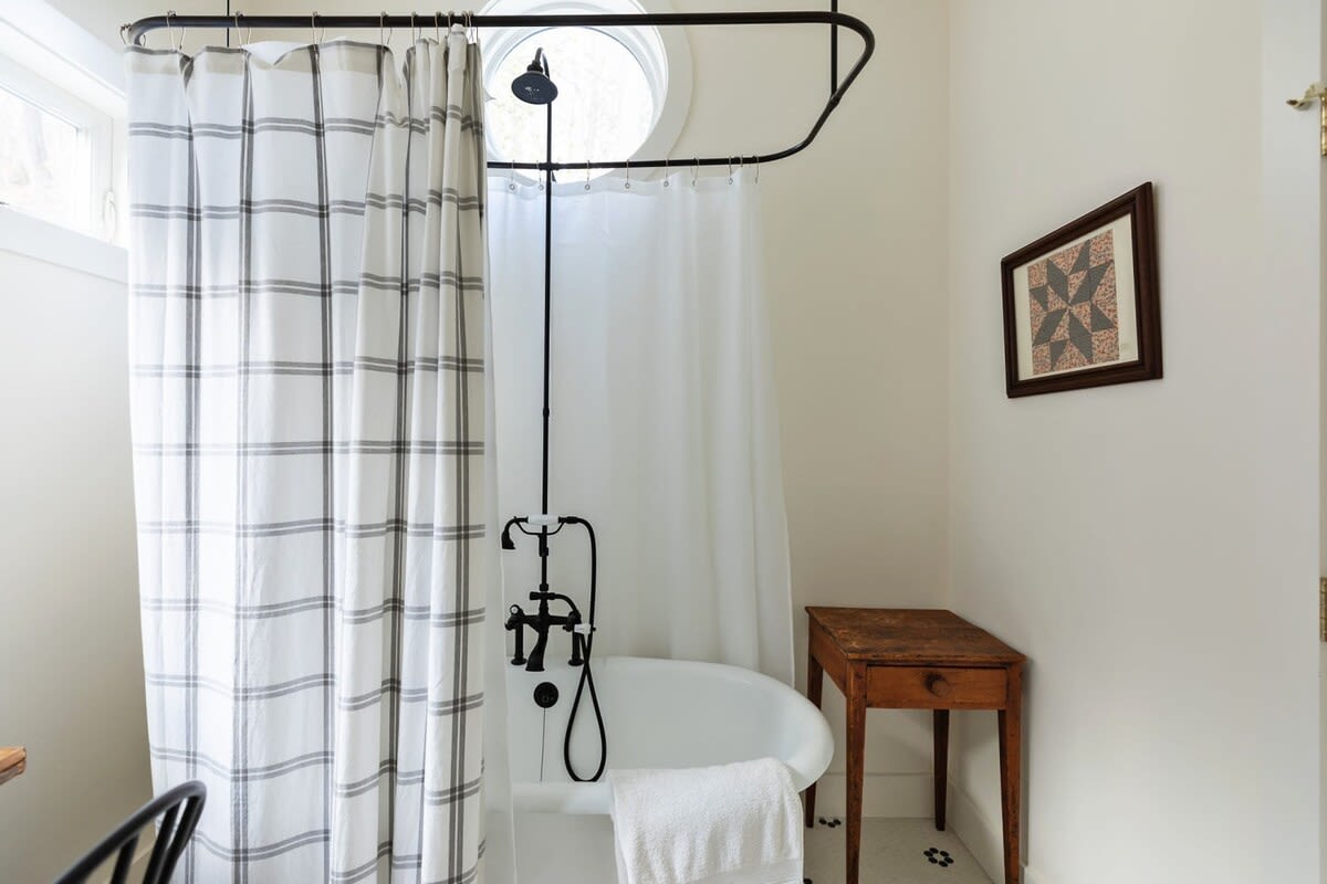 Charming bathroom with a white clawfoot tub, black shower fixtures, and a white shower curtain on a black rod. A striped shower curtain provides privacy, and a small rustic wooden table sits beside the tub.