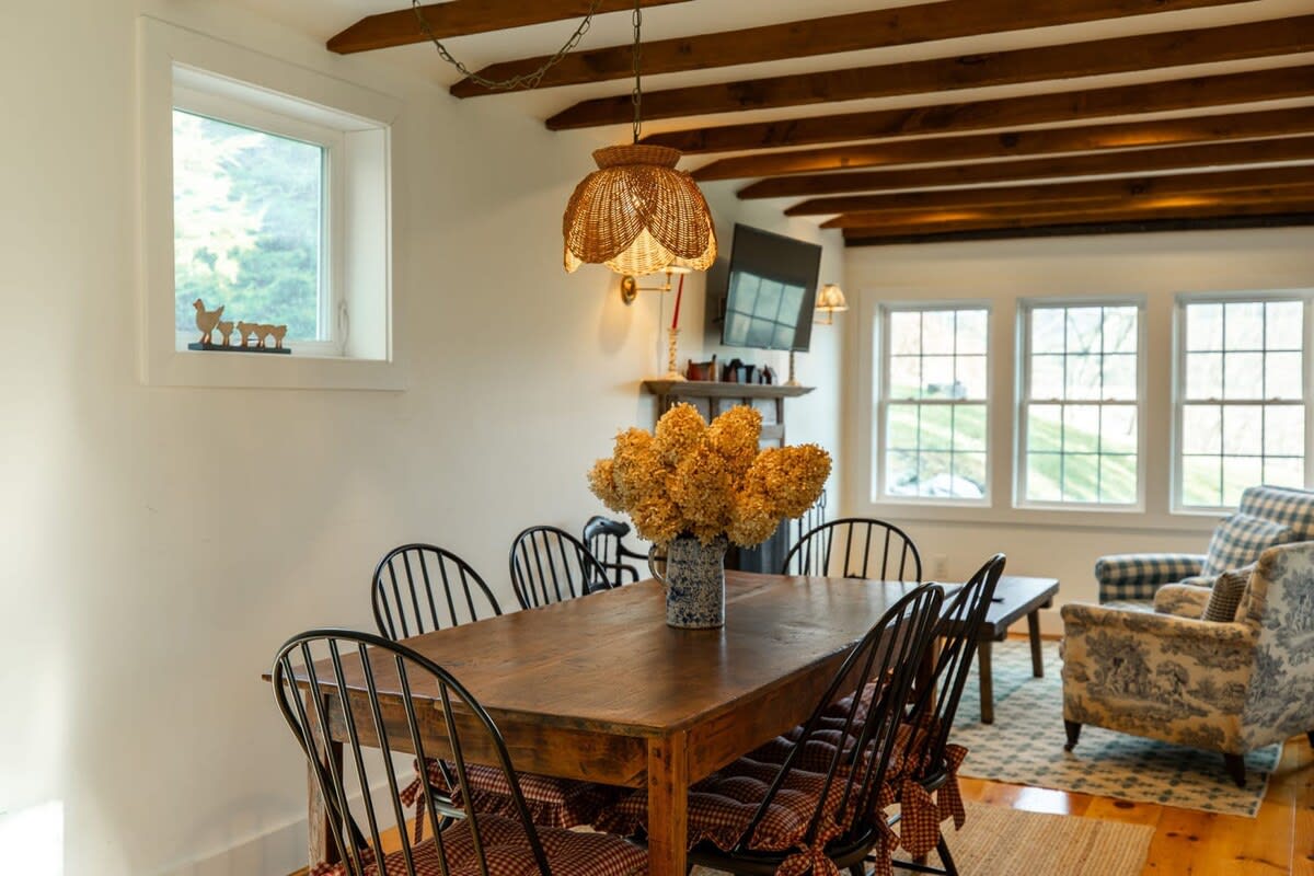A cozy dining area features a rustic wooden table and six black Windsor chairs with checkered cushions, illuminated by a woven pendant light. Exposed wooden beams cross the ceiling, and large windows offer views of the outdoors.