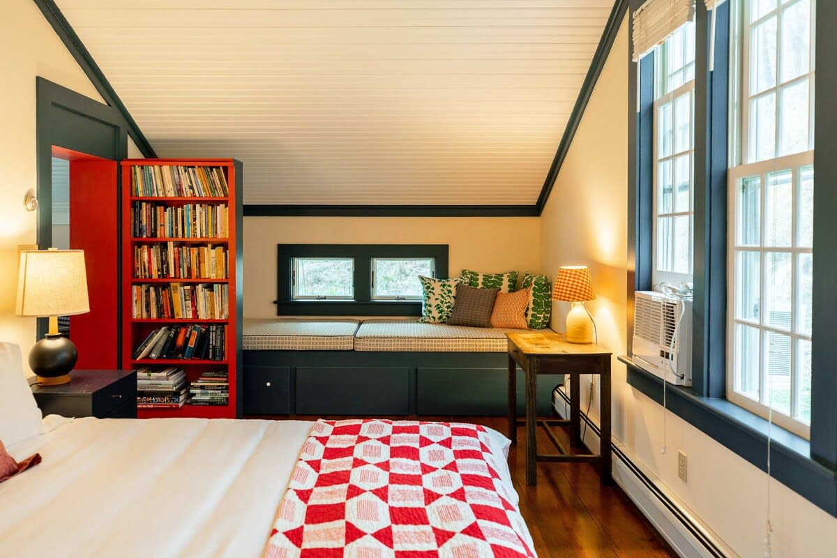 A bright bedroom features a bed with a red and white quilt, facing a built-in window seat with patterned cushions and throw pillows. A red bookshelf is visible to the left, and large windows with dark trim offer natural light.