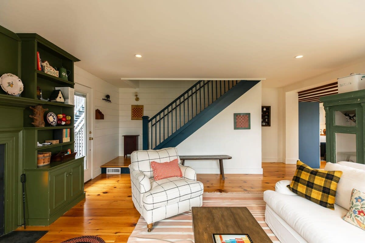 The spacious living room features a cozy fireplace with green built-in shelving, leading to a staircase with dark blue banisters. White walls and warm hardwood floors create a bright and welcoming space.