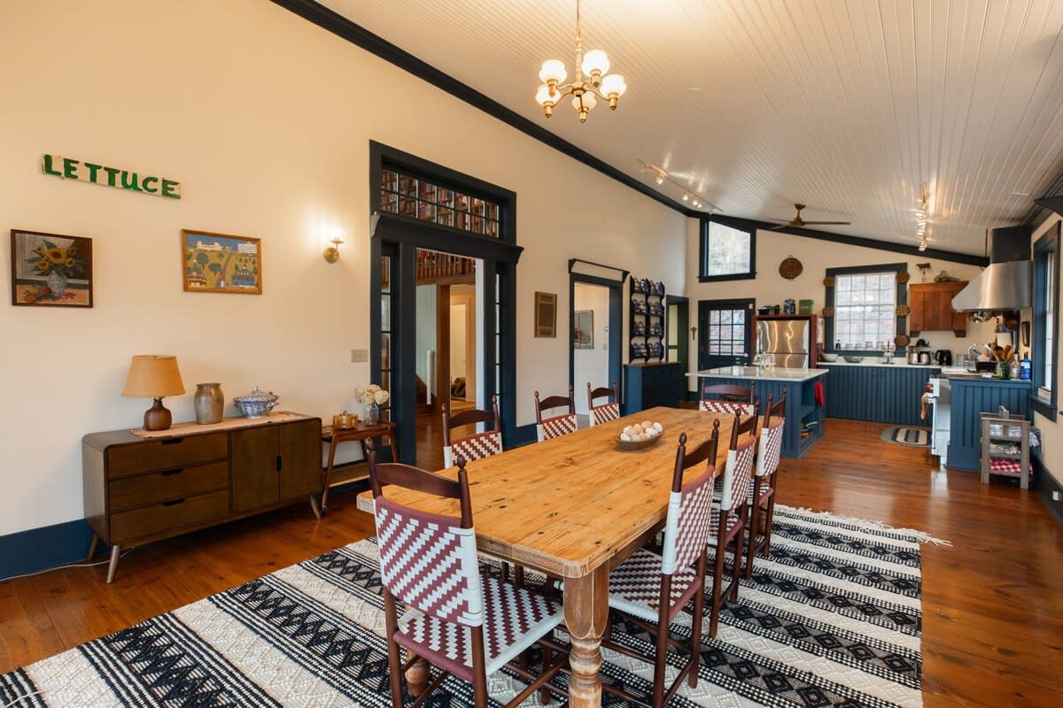 The dining room, with its long wooden table and woven-back chairs, flows into a modern kitchen featuring dark blue cabinetry and a large island. A "LETTUCE" sign adds a quirky touch to the light-colored walls.