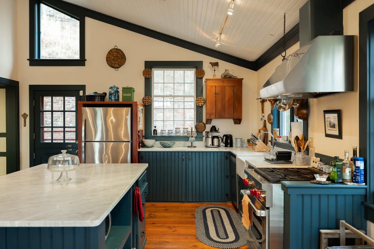 This kitchen view showcases dark blue beadboard cabinets, a large stainless steel refrigerator, and a farmhouse sink with a window above. Copper pots hang near the range hood, adding to the rustic-chic aesthetic.