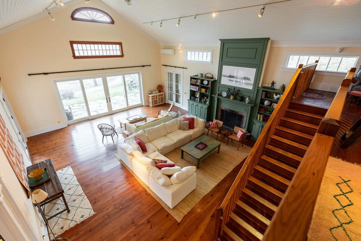 An expansive living room with vaulted ceilings and exposed beams features a large white sectional sofa, a green built-in fireplace with bookshelves, and a wooden staircase leading to an upper level. Hardwood floors and a large rug define the seating area.
