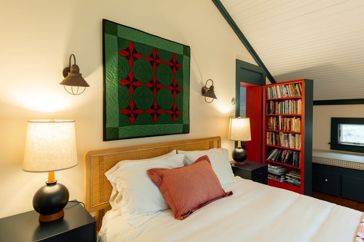 This bedroom showcases a bed with a cane headboard, white bedding, and a red throw pillow, flanked by dark nightstands with lamps. A vibrant red bookshelf filled with books is visible through an open doorway, adding a pop of color.