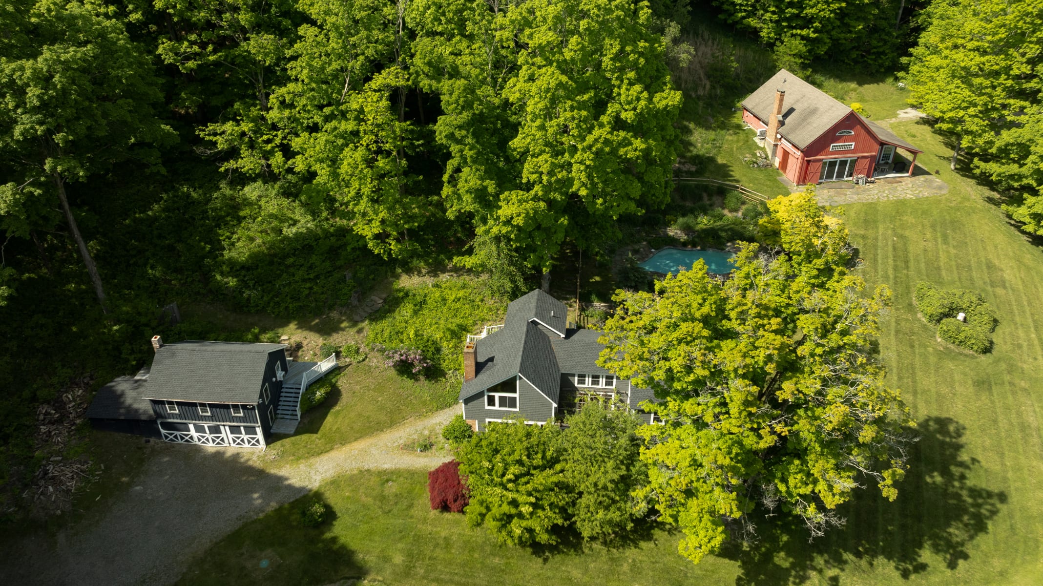 Wide aerial view of the property featuring a gray main house, a smaller dark gray guesthouse, and a red barn-like structure further up the hill. A blue-bottomed pool is visible near the red building, all surrounded by dense green foliage.