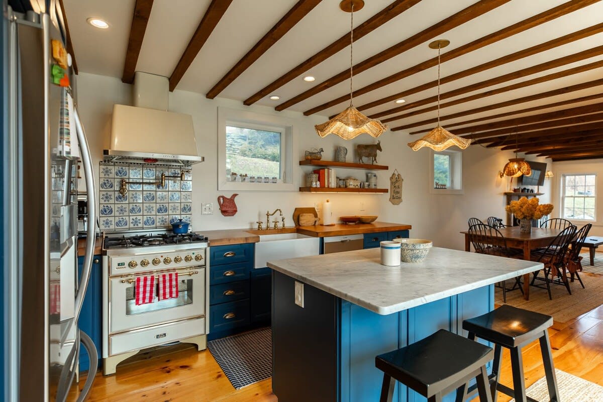 This charming kitchen features blue cabinetry, exposed wooden beams, and a marble-topped island with bar stools, leading into an open-concept dining area. A vintage-style range and farmhouse sink complete the rustic yet functional space.