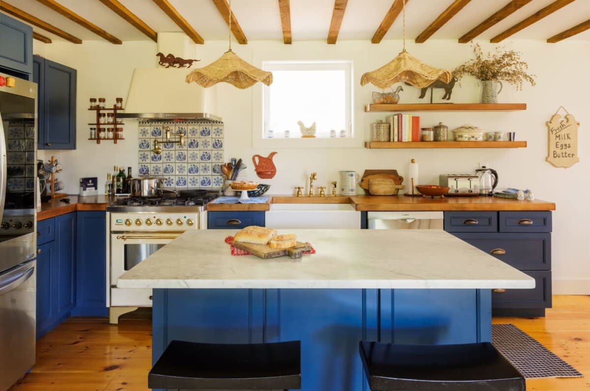 A rustic kitchen boasts exposed wooden ceiling beams and vibrant blue cabinetry, complemented by butcher block countertops and a white farmhouse sink. A marble-topped island with seating anchors the center, facing a vintage-style range with decorative tile backsplash.