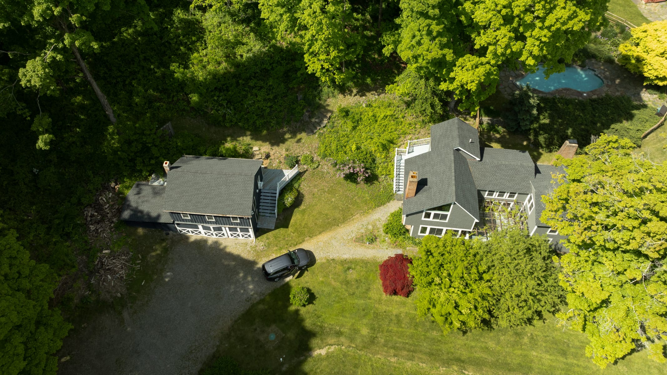 Aerial view showcasing a gray main house and a smaller dark gray guesthouse with white trim, connected by a gravel driveway. A black car is parked between the buildings, surrounded by vibrant green trees and lawn.