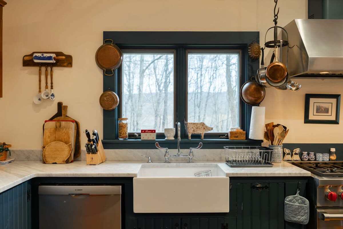 A detailed view of the kitchen sink area features a white farmhouse sink, light marble countertops, and a window overlooking a wooded landscape. Copper pots and various kitchen utensils are neatly arranged around the space.