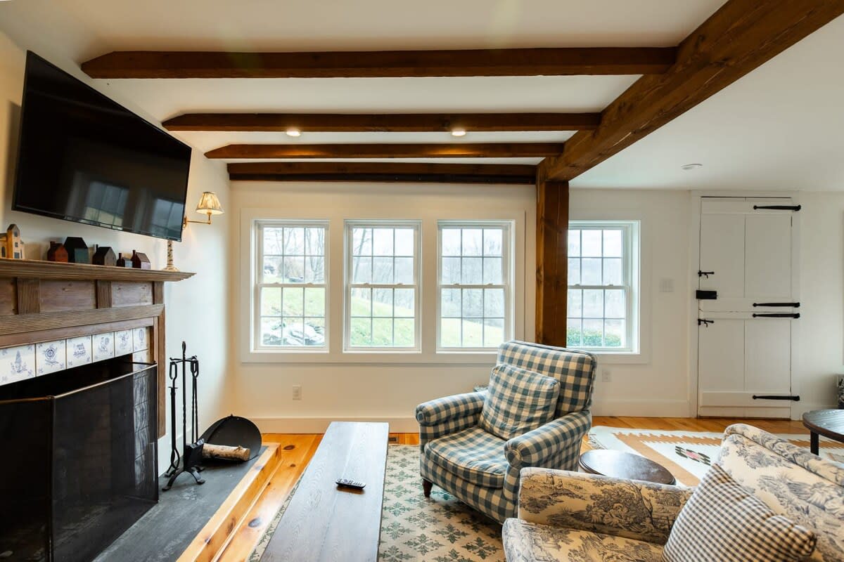 A comfortable living room features exposed wooden beams, light wood floors, and multiple windows overlooking a natural landscape. It includes a fireplace with a wooden mantel, a large flat-screen TV, and a plaid armchair.