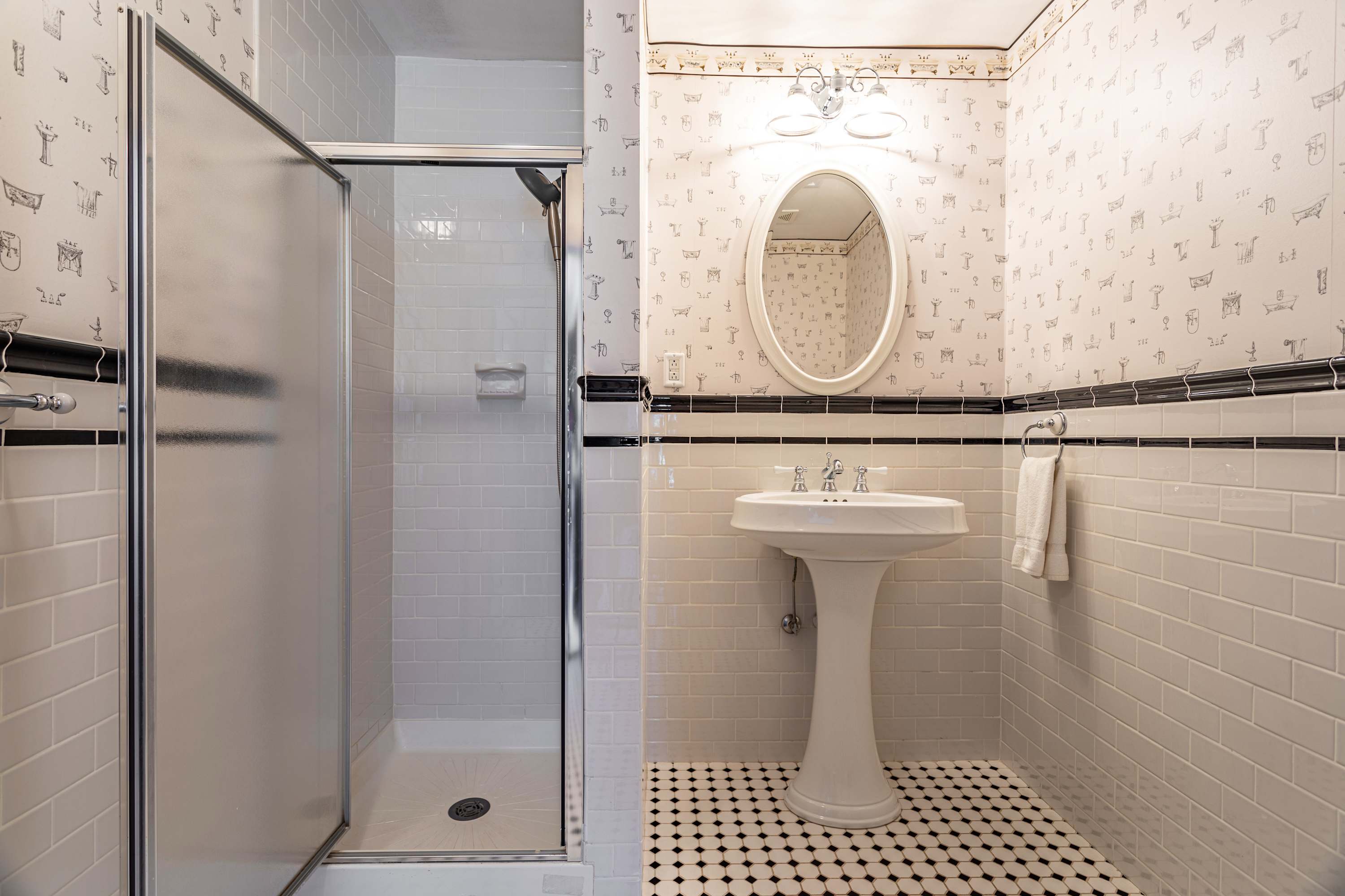 Vintage-style bathroom with white subway tiles, a glass-enclosed shower, and a pedestal sink, complemented by black and white patterned floor tiles and patterned wallpaper.