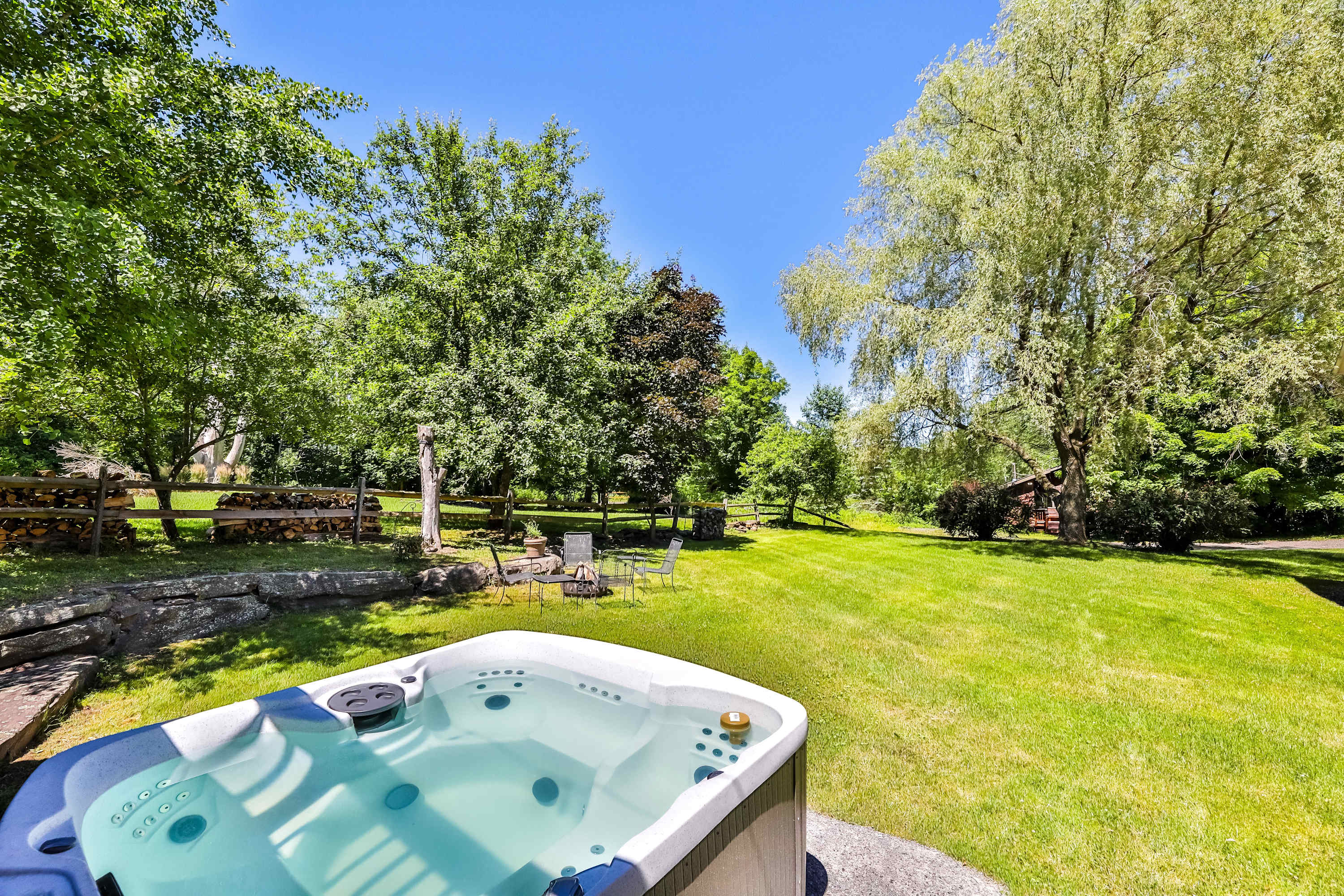 An outdoor hot tub with bubbling water is positioned on a stone patio in a lush green backyard, surrounded by mature trees and a wooden fence, with a fire pit area and seating in the distance.