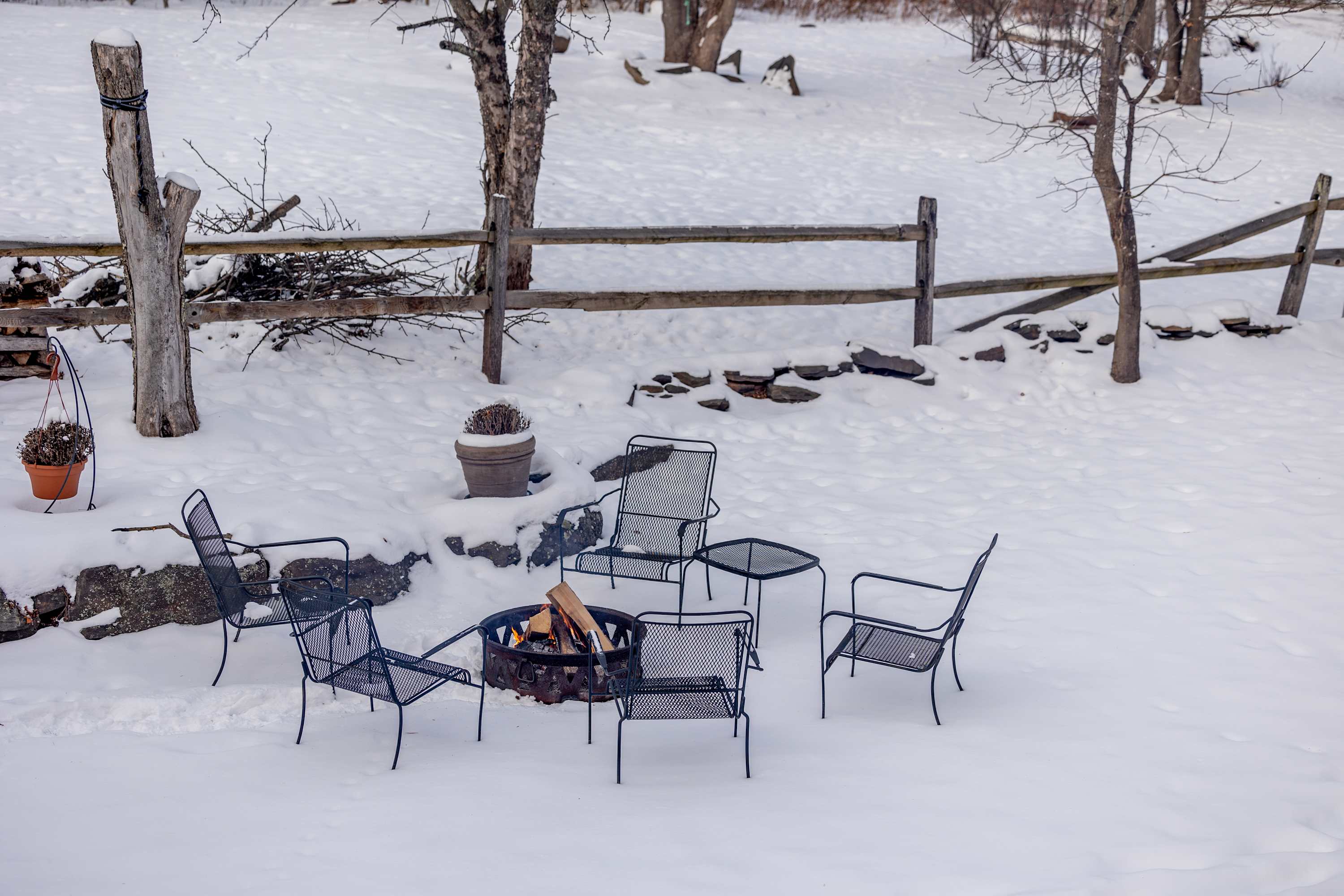 Outdoor fire pit area in a snow-covered yard, surrounded by several dark metal chairs, with a split-rail fence and bare trees in the background under a winter sky.
