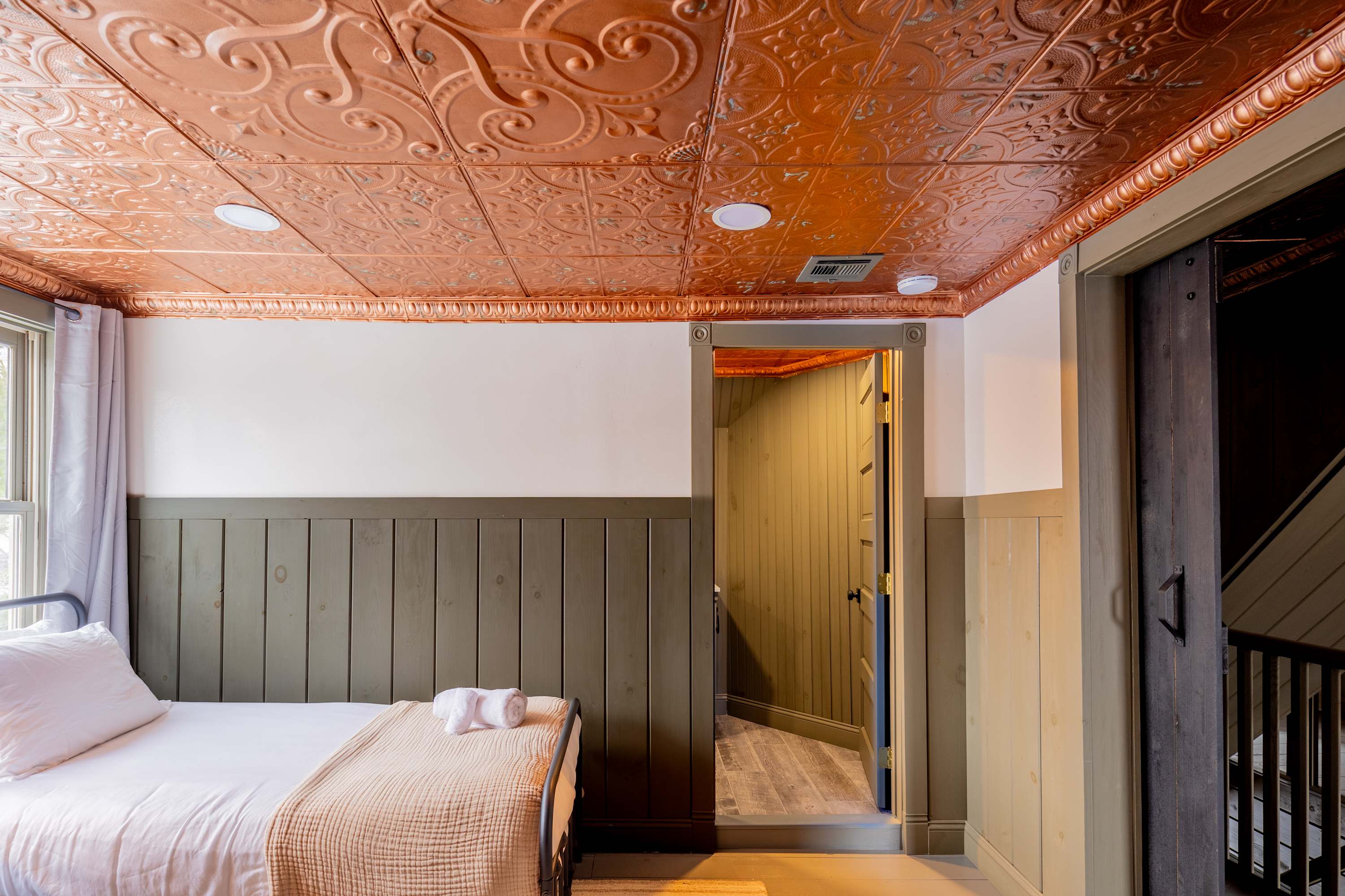 Bedroom with a black metal frame queen bed, light wood paneling on the lower walls, white upper walls, and a decorative copper-colored pressed tin ceiling.
