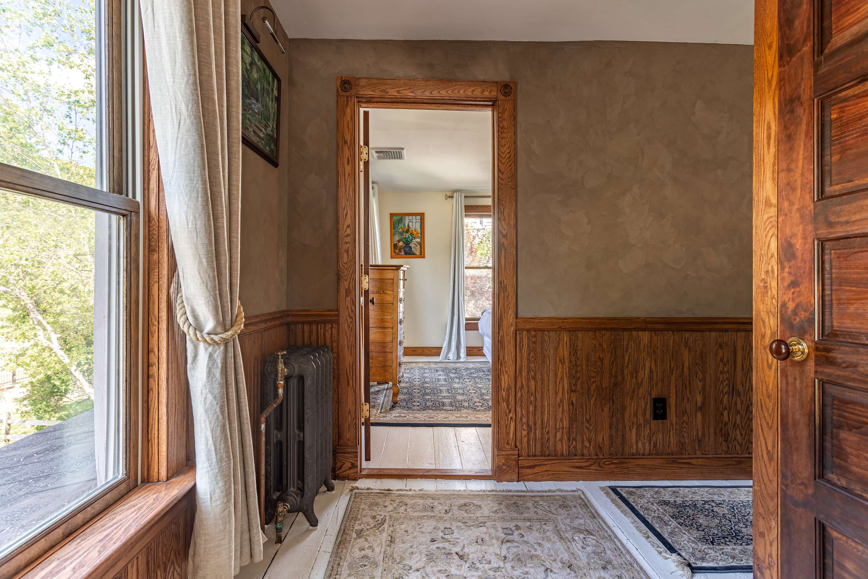 Bright interior space with white painted wooden floors and dark wood wainscoting, leading into another room through a wide wooden doorway. A vintage cast-iron radiator sits beneath a large window draped with natural-toned curtains.