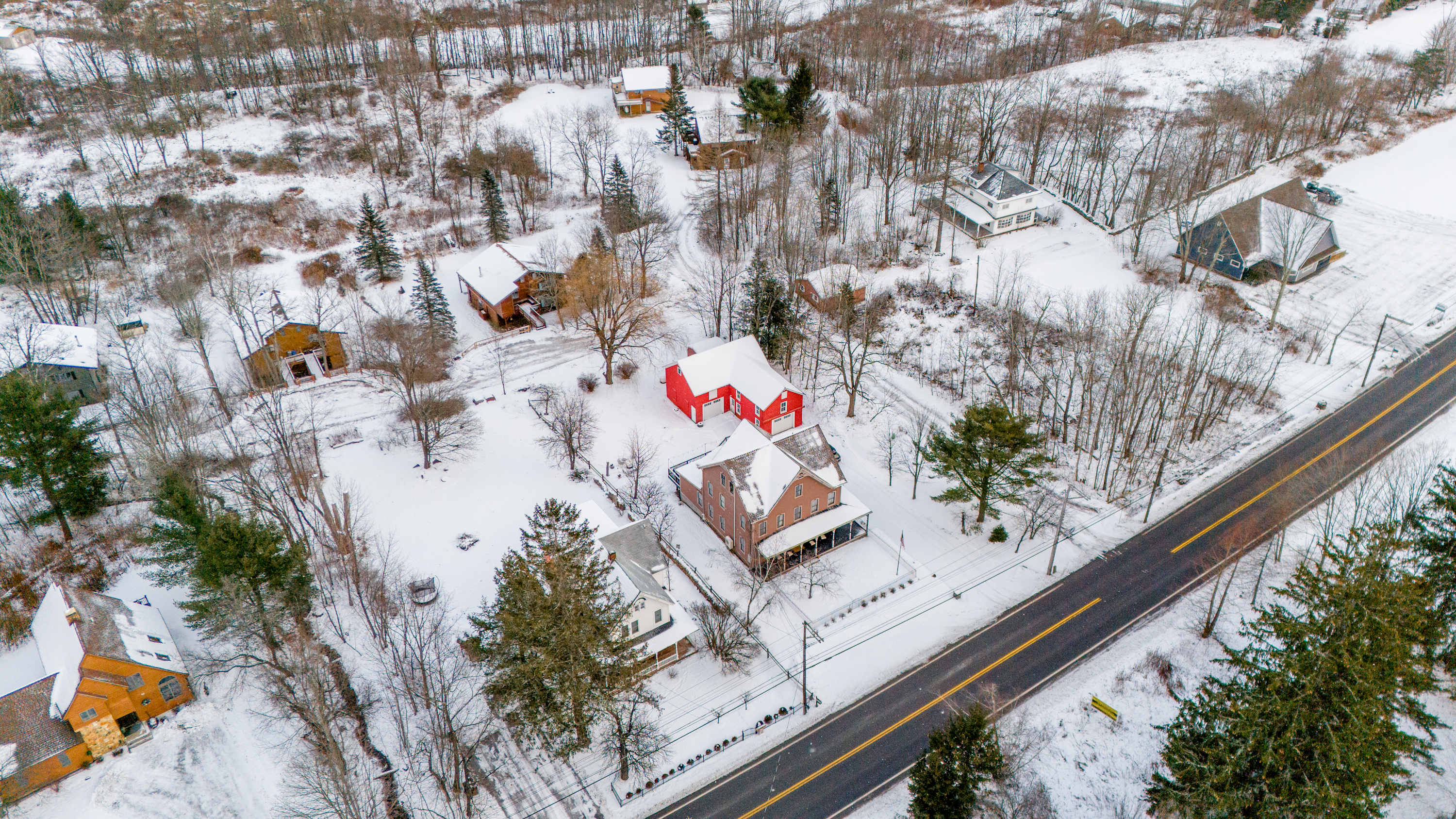 Aerial view of the property blanketed in snow, showcasing the main house with its wrap-around porch and the distinct red barn. A road runs alongside the property, surrounded by snow-dusted trees and other scattered buildings.